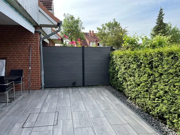 Fertige Terrasse aus grauen Platten in Holzoptik mit anthrazitfarbenem Sichtschutz und Splittstreifen entlang der Thuja-Hecke